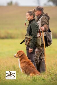 Kobieta stoi na łące z psem rasy Nova Scotia Duck Tolling Retriever przy lewej nodze. Obok niej stoi mężczyzna. Zdjęcie obrazuje trening dummy z Tollerem.